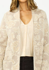 [Color: Taupe] A detail image of a brunette model wearing a light beige floral cardigan with an open front and patch pockets. 
