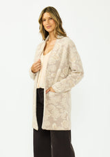 [Color: Taupe] A side facing image of a brunette model wearing a light beige floral cardigan with an open front and patch pockets. 