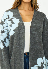 [Color: Charcoal] A detail image of a brunette woman wearing a dark gray open-front cardigan with large light blue floral patterns on the sleeves and lower front, worn over a white top and blue jeans .