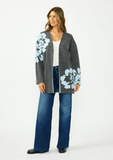 [Color: Charcoal] A front facing image of a brunette woman wearing a dark gray open-front cardigan with large light blue floral patterns on the sleeves and lower front, worn over a white top and blue jeans .