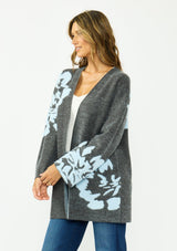 [Color: Charcoal] A side facing image of a brunette woman wearing a dark gray open-front cardigan with large light blue floral patterns on the sleeves and lower front, worn over a white top and blue jeans .
