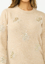 [Color: Heather Sand] A detail image of a brunette model wearing a light brown crew neck sweater with sequins and embellishment. 
