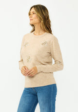 [Color: Heather Sand] A side facing image of a brunette model wearing a light brown crew neck sweater with sequins and embellishment. 