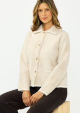 [Color: Natural/Taupe] A sitting image of a brunette model wearing a beige plaid sweater jacket with a fuzzy texture, collared neck, button front, and patch pockets. 