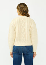 [Color: Natural] A back facing image of a brunette model wearing a cream cable knit sweater with a zip up front, a v neckline, and an orange yarn accent. Styled with a dark wash denim and beige loafers. 