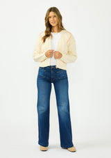 [Color: Natural] A front facing image of a brunette model wearing a cream cable knit sweater with a zip up front, a v neckline, and an orange yarn accent. Styled with a dark wash denim and beige loafers. 