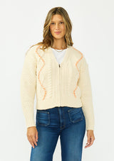 [Color: Natural] A front facing image of a brunette model wearing a cream cable knit sweater with a zip up front, a v neckline, and an orange yarn accent. 
