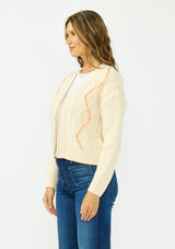 [Color: Natural] A side facing image of a brunette model wearing a cream cable knit sweater with a zip up front, a v neckline, and an orange yarn accent. Styled with a dark wash denim and beige loafers. 