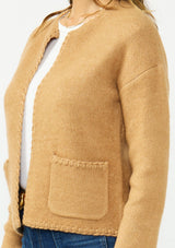 [Color: Camel] A detail image of a brunette model wearing a camel brown cardigan with an open front, patch pockets, and blanket stitch trims. Styled over a white tee and dark wash denim. 