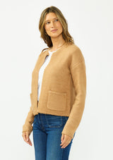 [Color: Camel] A side facing image of a brunette model wearing a camel brown cardigan with an open front, patch pockets, and blanket stitch trims. Styled over a white tee and dark wash denim. 