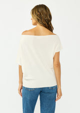 [Color: Natural] A back facing image a brunette model wearing an off white off the shoulder sweater top with short sleeves and a fitted ribbed hem. 