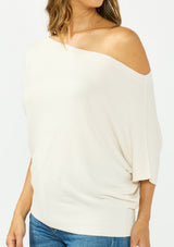[Color: Natural] A detail image a brunette model wearing an off white off the shoulder sweater top with short sleeves and a fitted ribbed hem. 