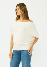 [Color: Natural] A side facing image a brunette model wearing an off white off the shoulder sweater top with short sleeves and a fitted ribbed hem. 