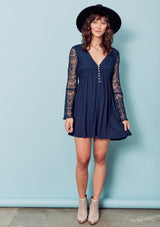 [Color: Dark Navy] A brunette woman wearing a blue sheer lace mini dress. Featuring a feminine v neckline, a button front detail, an empire waist, long sheer lace bell sleeves, and a sexy sheer lace back detail.