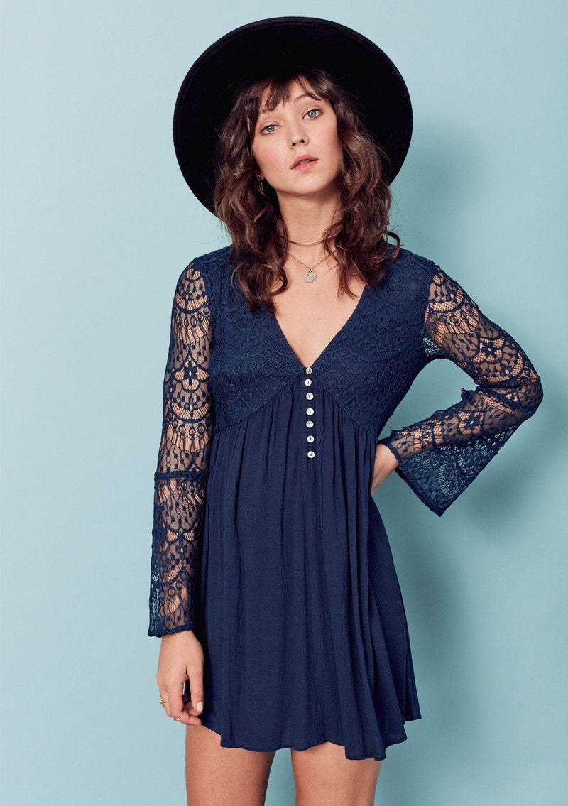 [Color: Dark Navy] A brunette woman wearing a blue sheer lace mini dress. Featuring a feminine v neckline, a button front detail, an empire waist, long sheer lace bell sleeves, and a sexy sheer lace back detail.