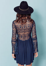 [Color: Dark Navy] A brunette woman wearing a blue sheer lace mini dress. Featuring a feminine v neckline, a button front detail, an empire waist, long sheer lace bell sleeves, and a sexy sheer lace back detail.