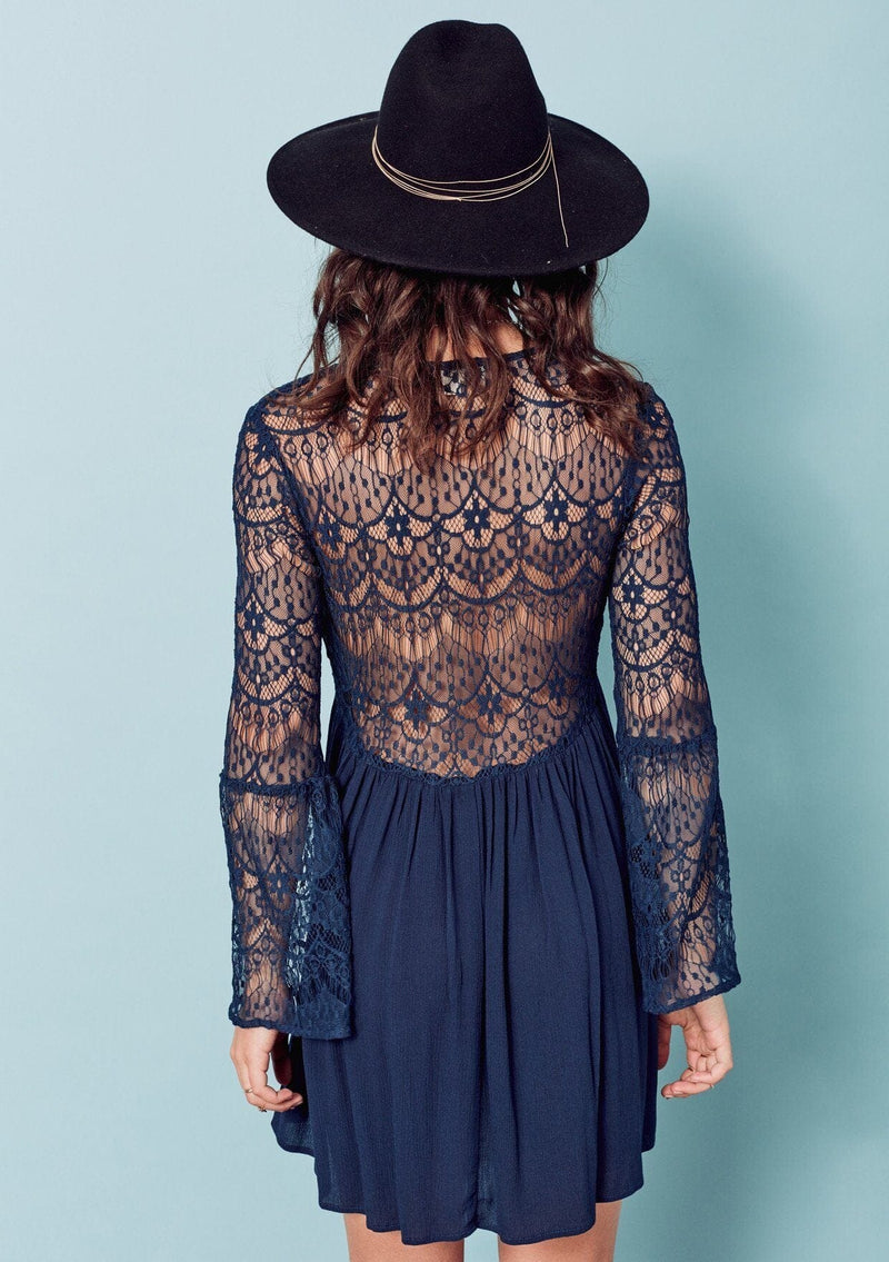 [Color: Dark Navy] A brunette woman wearing a blue sheer lace mini dress. Featuring a feminine v neckline, a button front detail, an empire waist, long sheer lace bell sleeves, and a sexy sheer lace back detail.
