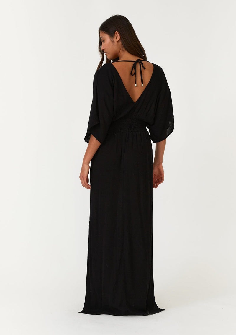 [Color: Black] A back facing image of a brunette model wearing a resort ready black maxi dress. With half length kimono sleeves, a plunging v neckline, a smocked elastic empire waist, side slits, and an open back with tie closure.