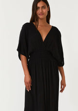 [Color: Black] A close up front facing image of a brunette model wearing a resort ready black maxi dress. With half length kimono sleeves, a plunging v neckline, a smocked elastic empire waist, side slits, and an open back with tie closure.