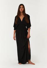 [Color: Black] A front facing image of a brunette model wearing a resort ready black maxi dress. With half length kimono sleeves, a plunging v neckline, a smocked elastic empire waist, side slits, and an open back with tie closure.