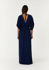 [Color: Pacific Blue] A back facing image of a brunette model wearing a resort ready blue maxi dress. With half length kimono sleeves, a plunging v neckline, a smocked elastic empire waist, side slits, and an open back with tie closure.