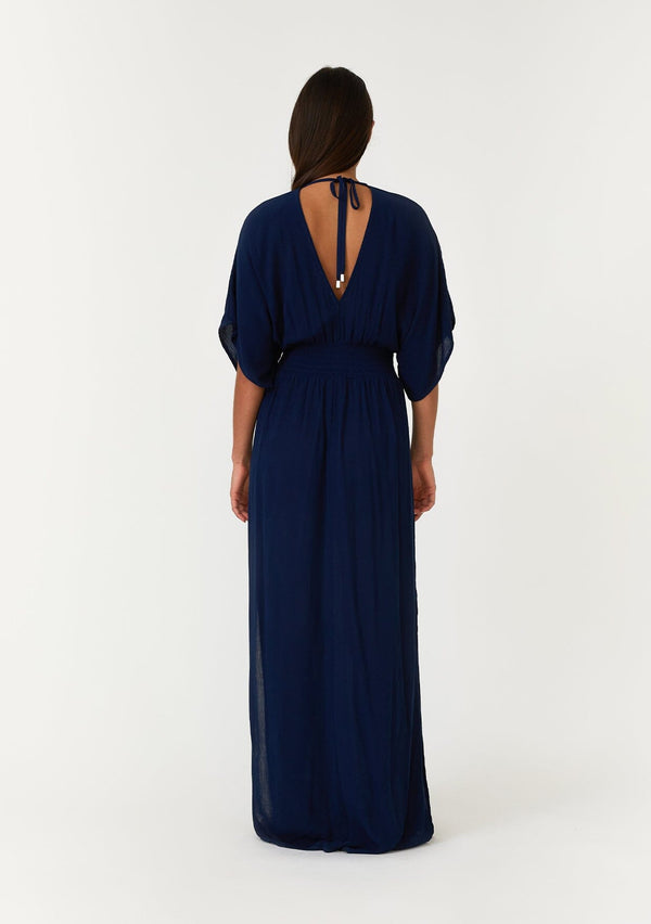 [Color: Pacific Blue] A back facing image of a brunette model wearing a resort ready blue maxi dress. With half length kimono sleeves, a plunging v neckline, a smocked elastic empire waist, side slits, and an open back with tie closure.