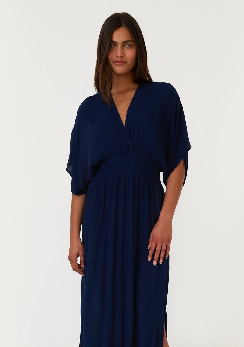 [Color: Pacific Blue] A close up front facing image of a brunette model wearing a resort ready blue maxi dress. With half length kimono sleeves, a plunging v neckline, a smocked elastic empire waist, side slits, and an open back with tie closure.