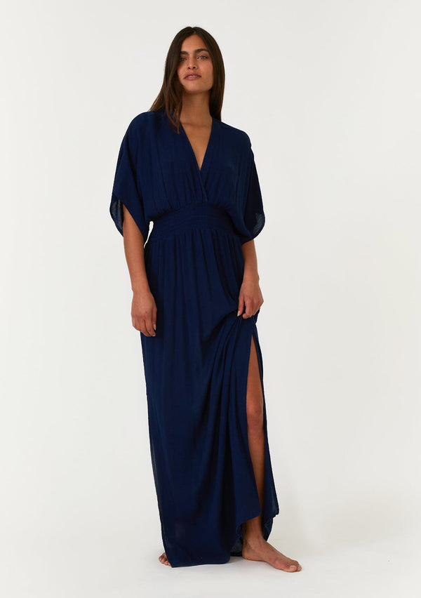 [Color: Pacific Blue] A front facing image of a brunette model wearing a resort ready blue maxi dress. With half length kimono sleeves, a plunging v neckline, a smocked elastic empire waist, side slits, and an open back with tie closure.