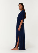 [Color: Pacific Blue] A side facing image of a brunette model wearing a resort ready blue maxi dress. With half length kimono sleeves, a plunging v neckline, a smocked elastic empire waist, side slits, and an open back with tie closure.