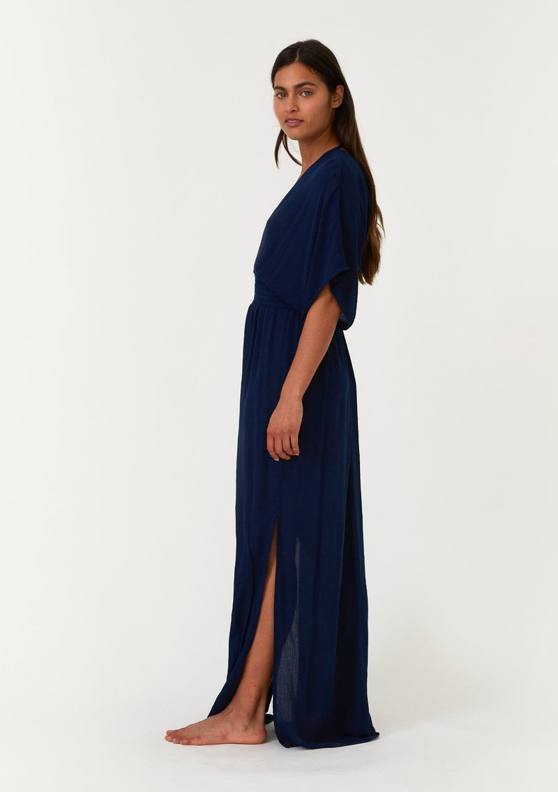 [Color: Pacific Blue] A side facing image of a brunette model wearing a resort ready blue maxi dress. With half length kimono sleeves, a plunging v neckline, a smocked elastic empire waist, side slits, and an open back with tie closure.