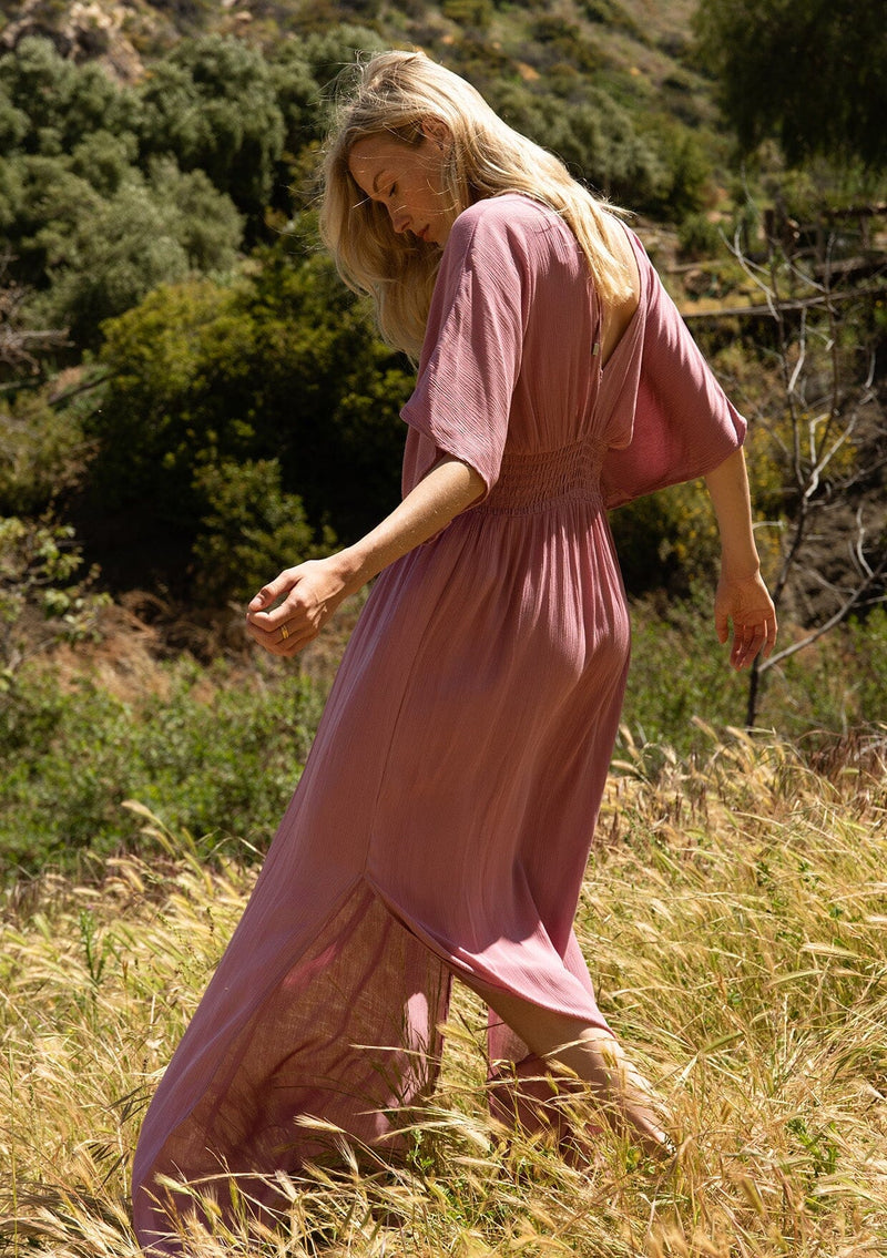 [Color: Smokey Orchid] A back facing image of a blonde model wearing a resort ready light purple maxi dress. With half length kimono sleeves, a plunging v neckline, a smocked elastic empire waist, side slits, and an open back with tie closure.