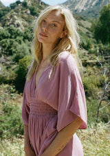 [Color: Smokey Orchid] A close up side facing image of a blonde model wearing a resort ready light purple maxi dress. With half length kimono sleeves, a plunging v neckline, a smocked elastic empire waist, side slits, and an open back with tie closure.