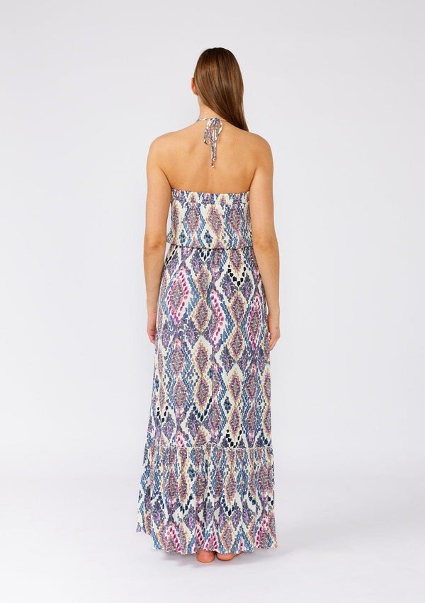 [Color: Natural/Dusty Blue] A back facing image of a brunette model wearing a stunning sleeveless beach maxi dress designed in a blue bohemian print. With a halter neckline and adjustable tie at the back neckline, an elastic waist, and a long flowy skirt with a ruffle trimmed tiered hemline. 