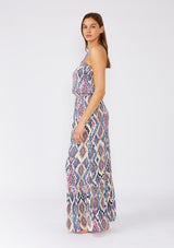 [Color: Natural/Dusty Blue] A side facing image of a brunette model wearing a stunning sleeveless beach maxi dress designed in a blue bohemian print. With a halter neckline and adjustable tie at the back neckline, an elastic waist, and a long flowy skirt with a ruffle trimmed tiered hemline. 