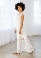 [Color: Off White] A side facing image of a brunette woman wearing a white clip dot chiffon maxi dress with a sleeveless design, a v neckline, and a tie front detail. 