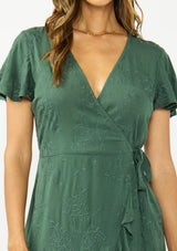 [Color: Forest Green] A detail image of a brunette model wearing a green embroidered wrap dress with short sleeves, a ruffle trimmed hem, and a side tie detail. 