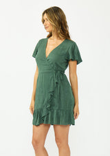 [Color: Forest Green] A side facing image of a brunette model wearing a green embroidered wrap dress with short sleeves, a ruffle trimmed hem, and a side tie detail. 