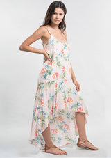 [Color: Blush/Green] A side facing image of a brunette model wearing a tropical floral print maxi dress with a high low hemline, a scoop neckline, and spaghetti straps. 