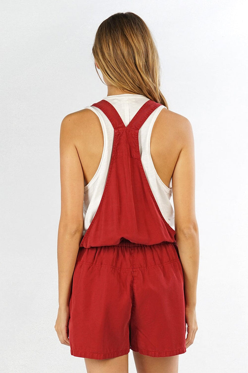 [Color: Brick] A back facing image of a blonde model wearing a red overall romper over a tank top with open sides, a front zipper details, drawstring waist, and side pockets. 