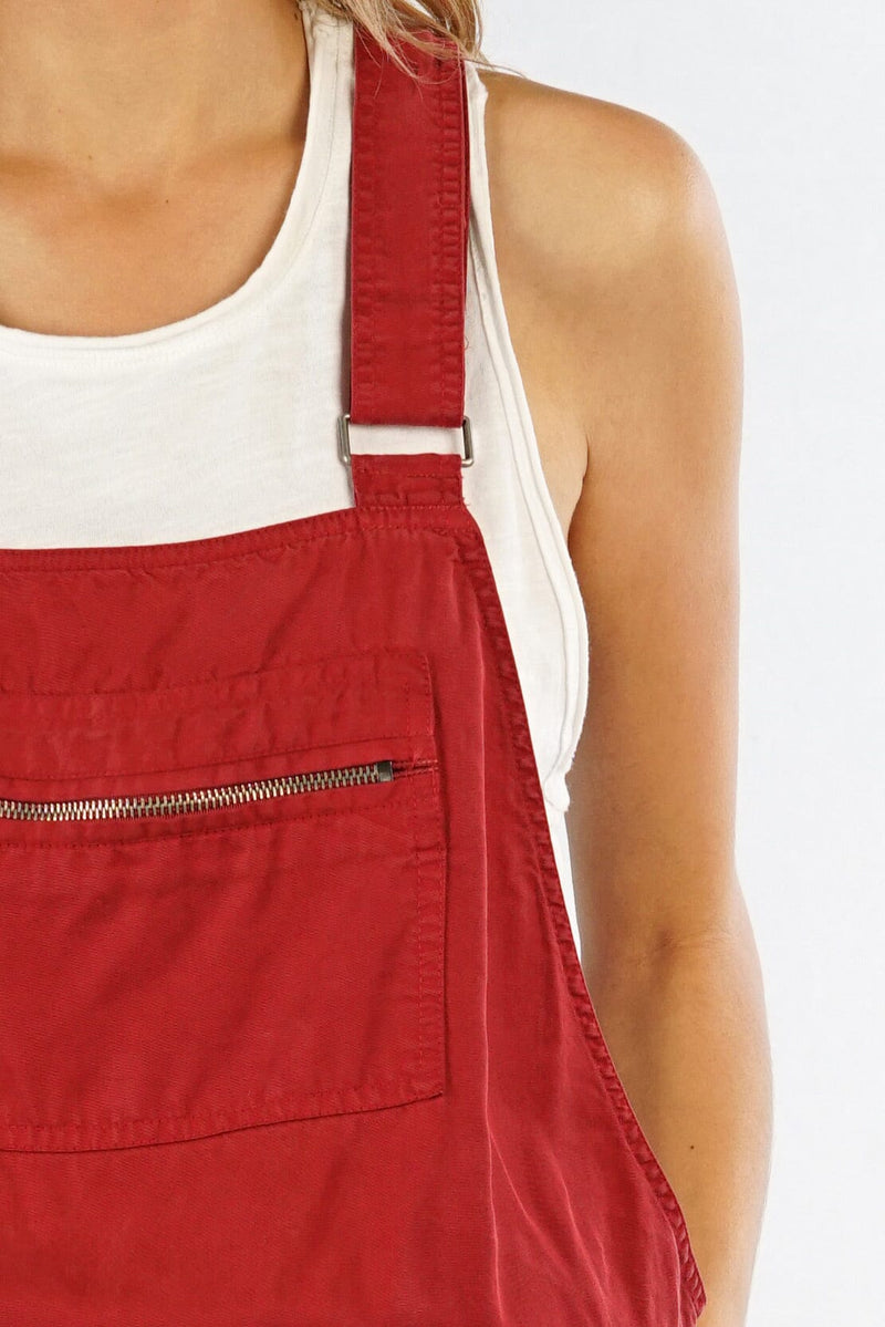 [Color: Brick] A detail image of a blonde model wearing a red overall romper over a tank top with open sides, a front zipper details, drawstring waist, and side pockets. 
