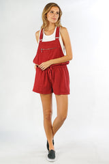 [Color: Brick] A front facing image of a blonde model wearing a red overall romper over a tank top with open sides, a front zipper details, drawstring waist, and side pockets. 