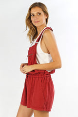 [Color: Brick] A side facing image of a blonde model wearing a red overall romper over a tank top with open sides, a front zipper details, drawstring waist, and side pockets. 