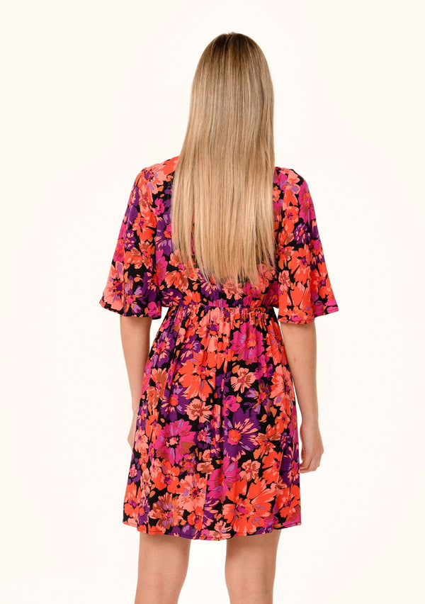 [Color: Black/Fuchsia] A back facing image of a blonde model wearing a flowy fall mini dress in a bright pink floral print. With half length short sleeves, a deep v neckline, and an empire waist. 