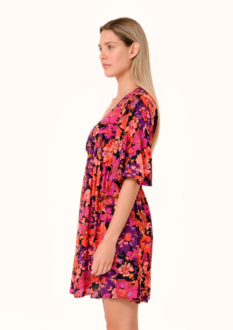 [Color: Black/Fuchsia] A side facing image of a blonde model wearing a flowy fall mini dress in a bright pink floral print. With half length short sleeves, a deep v neckline, and an empire waist. 