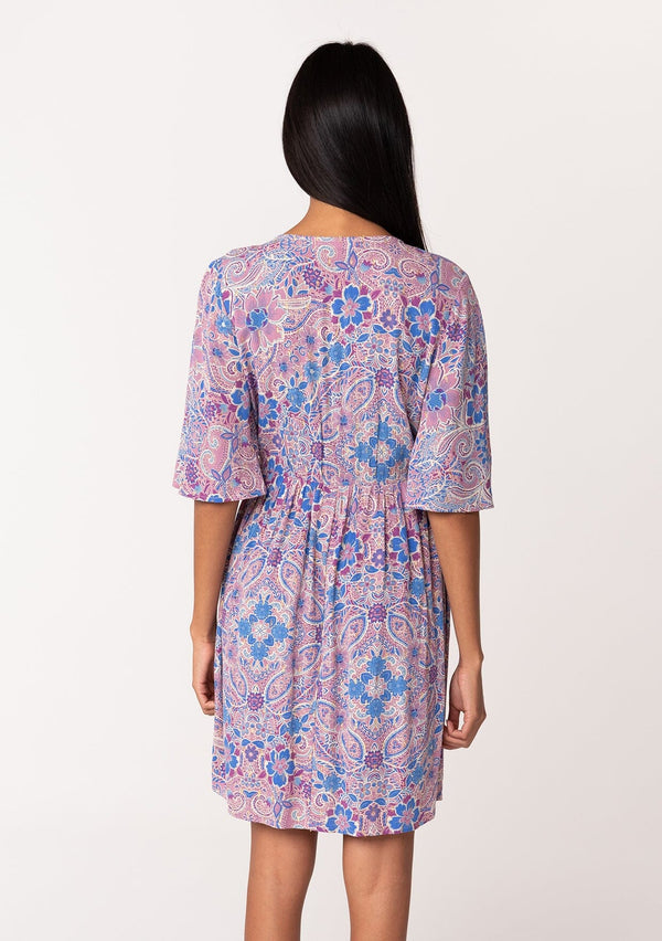 [Color: Dusty Rose/Blue] A back facing image of a brunette model wearing a flowy summer mini dress with half length sleeves, a v neckline, and an empire waist. Designed in a pink and blue floral print with gold clip dot details throughout. 