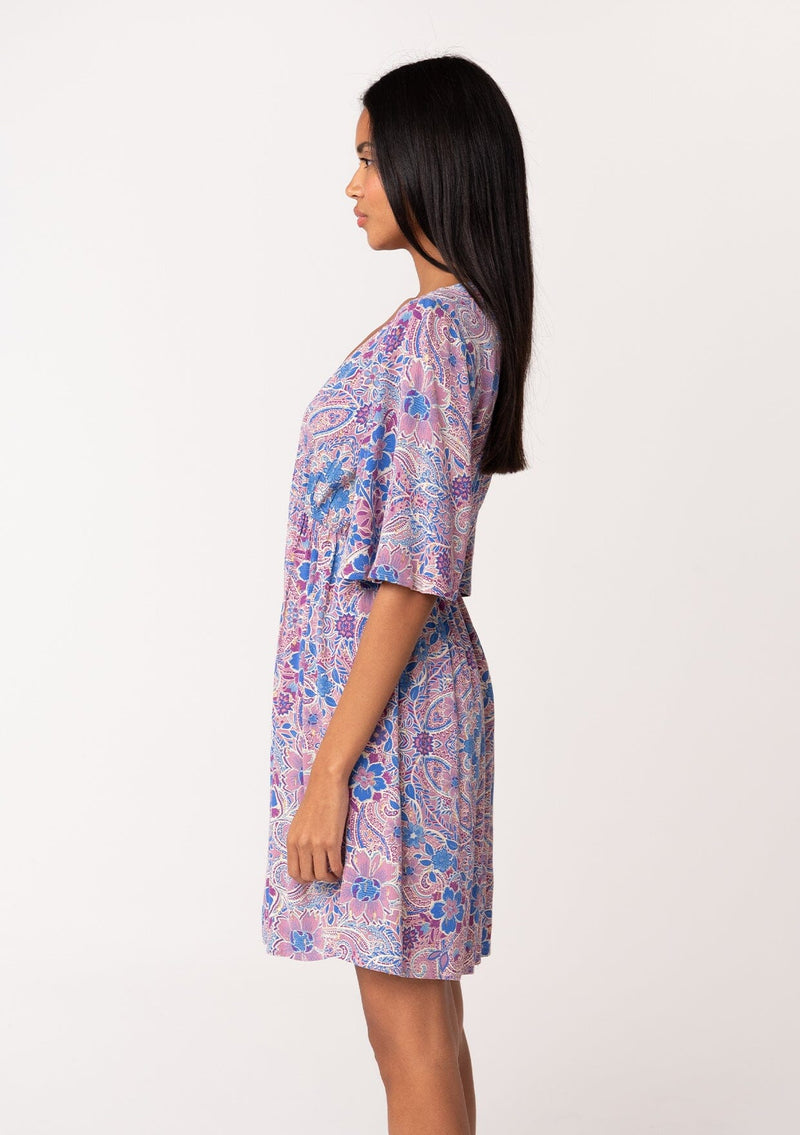 [Color: Dusty Rose/Blue] A side facing image of a brunette model wearing a flowy summer mini dress with half length sleeves, a v neckline, and an empire waist. Designed in a pink and blue floral print with gold clip dot details throughout. 