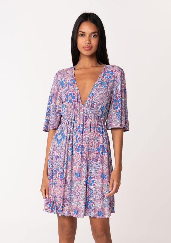 [Color: Dusty Rose/Blue] A front facing image of a brunette model wearing a flowy summer mini dress with half length sleeves, a v neckline, and an empire waist. Designed in a pink and blue floral print with gold clip dot details throughout. 