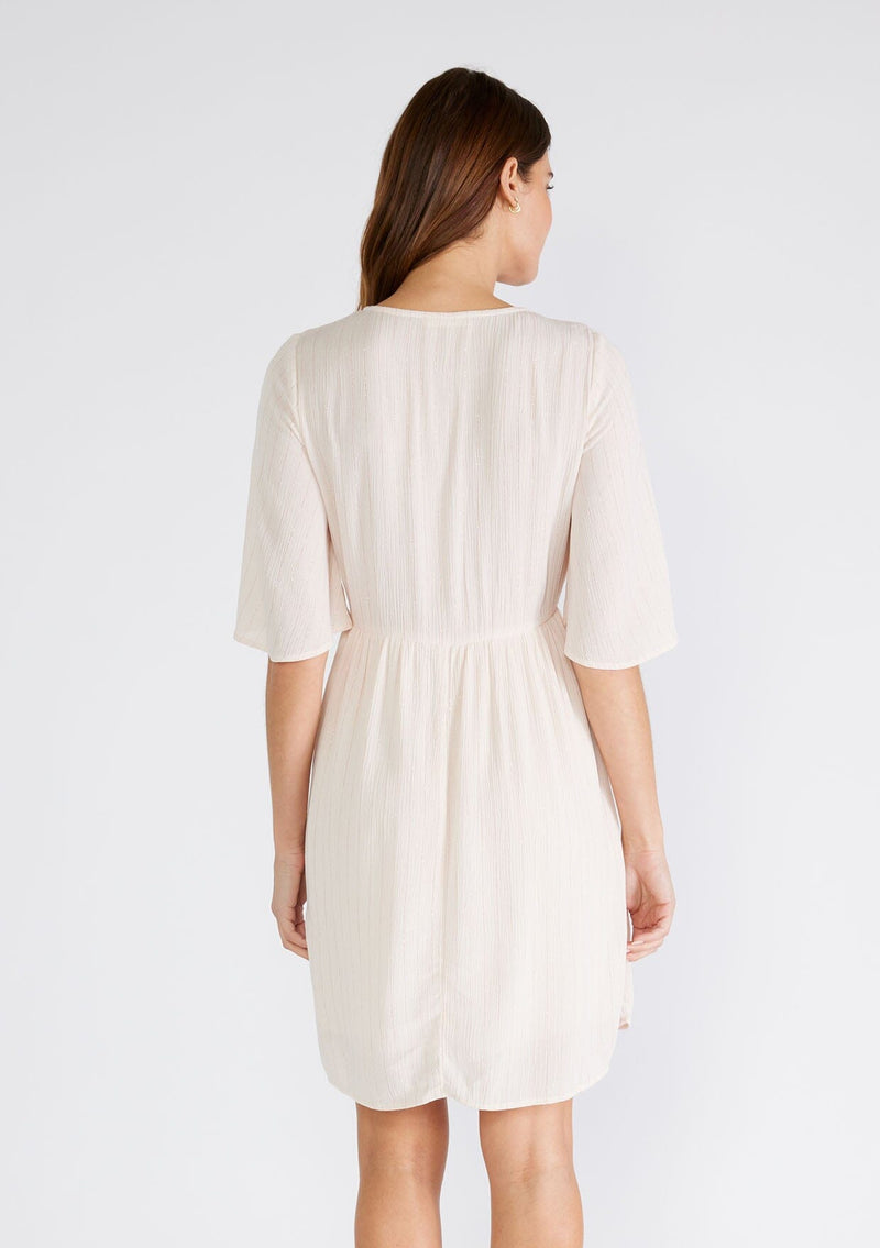 [Color: Light Peach] A back facing image of a brunette model wearing a classic flowy mini dress in light peach with a gold metallic thread detail. With short half length sleeves, a v neckline, and an empire waist.