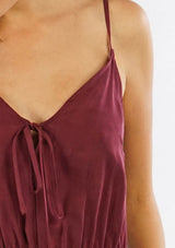 [Color: Burgundy] A detail image of a woman wearing a one piece jumpsuit. Featuring a wide leg, a front keyhole detail, an elastic cinched waist, and side pockets.