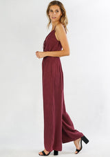 [Color: Burgundy] A side facing image of a woman wearing a one piece jumpsuit. Featuring a wide leg, a front keyhole detail, an elastic cinched waist, and side pockets.